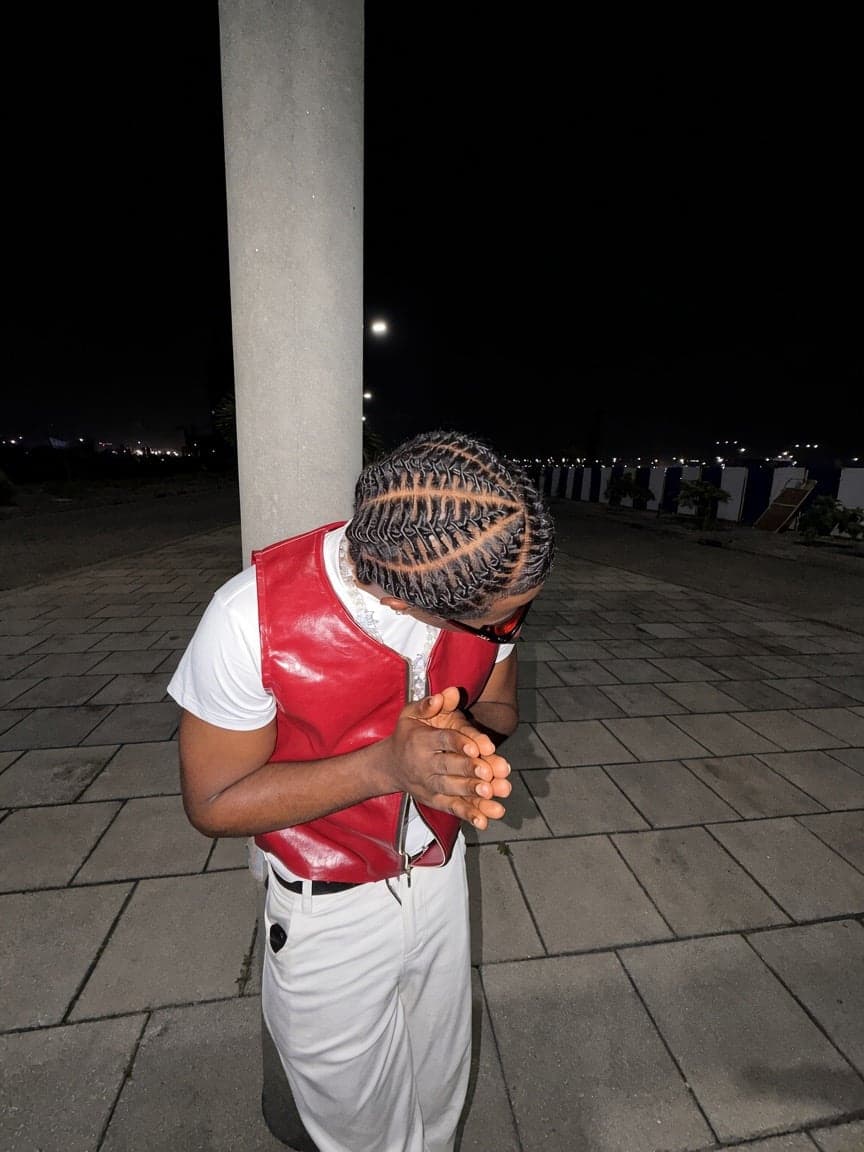 Head bowed showing stitched braids, red leather vest and white trousers at night
