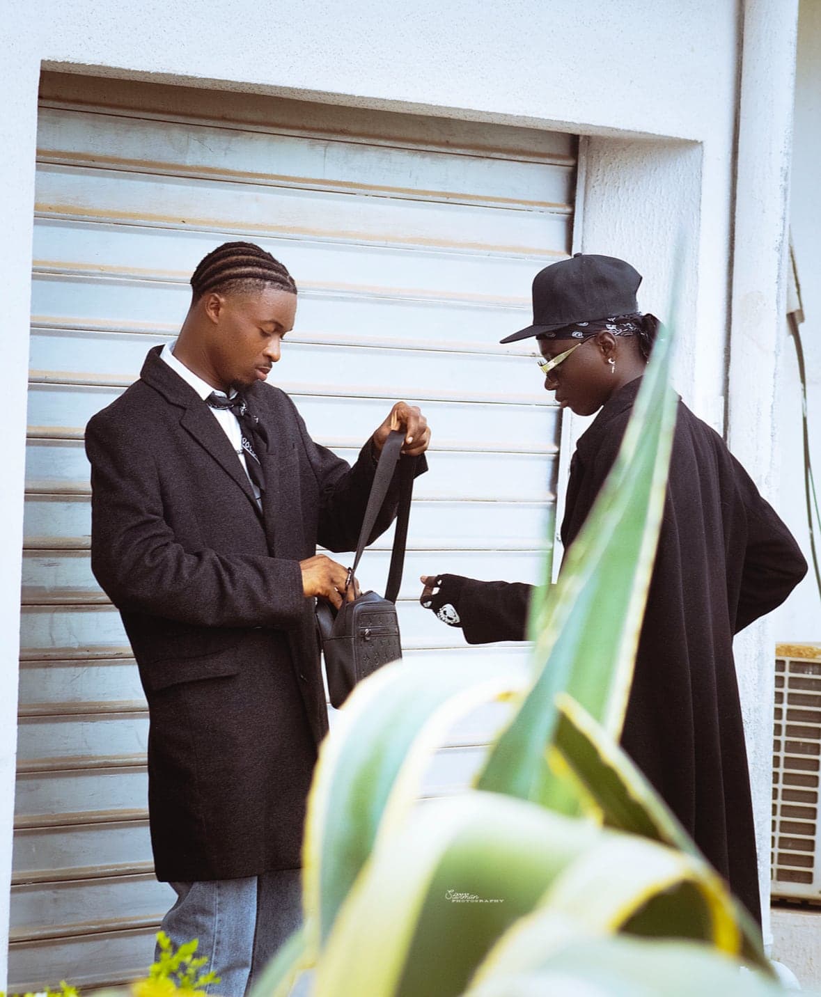 Two models in black coats passing a crossbody bag in front of a shutter