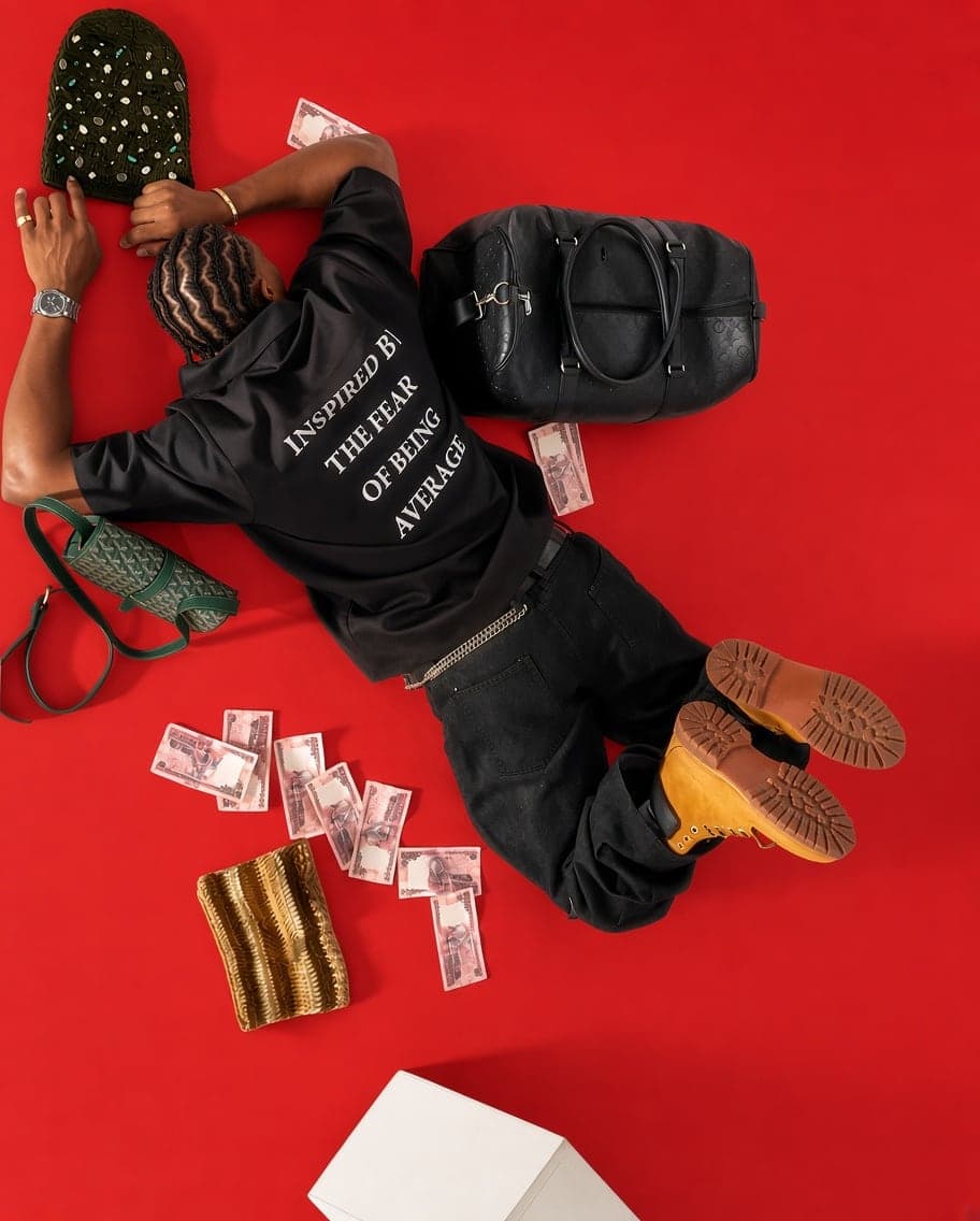 Lying on a red floor in a black tee reading Inspired by the fear of being average, surrounded by bags and banknotes