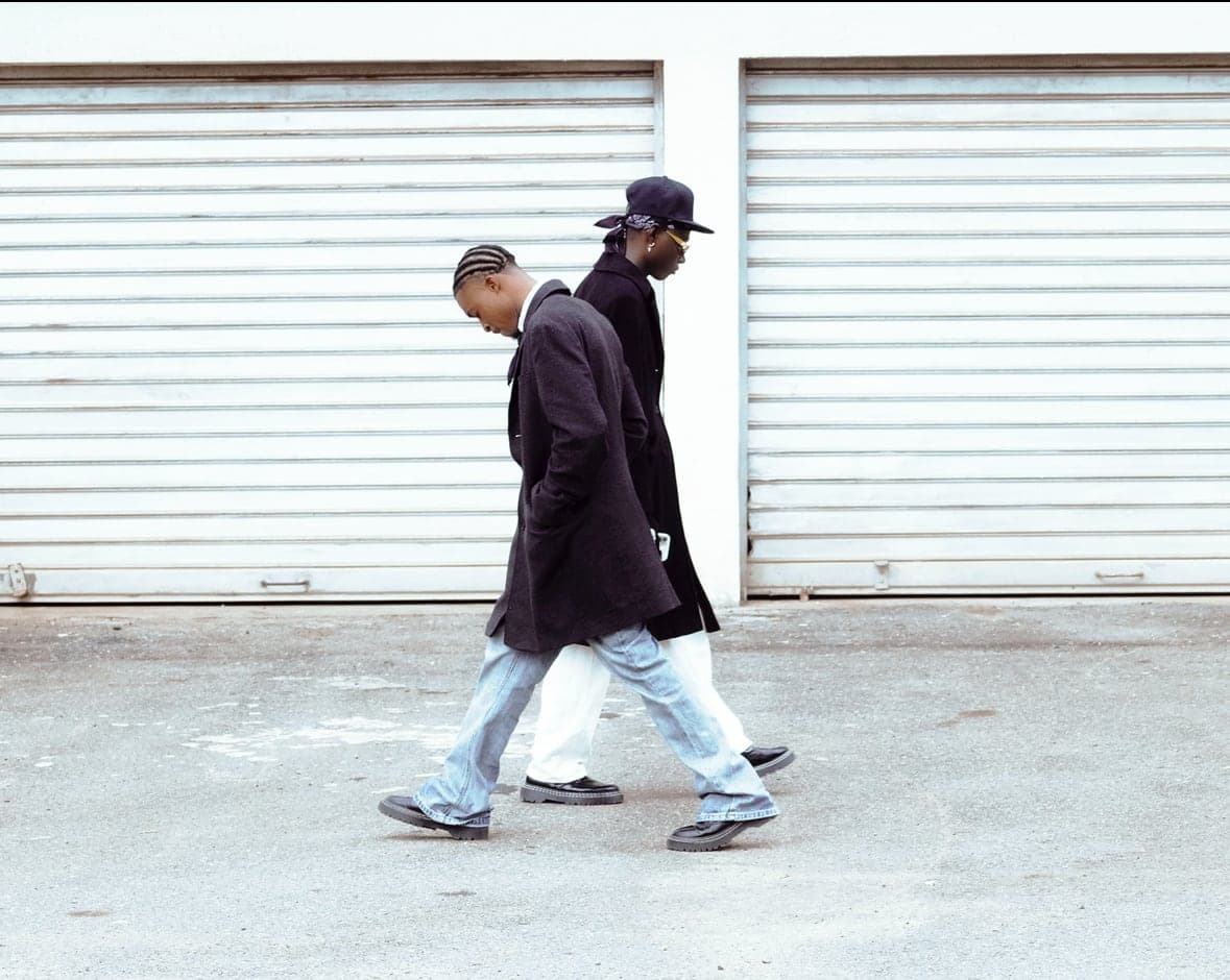 Two models in long black coats walking past white shutters