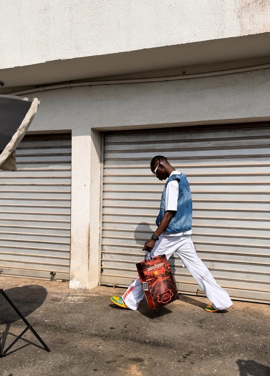 Walking past shutters in a denim vest and white trousers, carrying a red bag